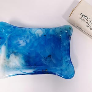 May include: A blue and white marbled soap dish with rounded edges, next to a bar of soap. The soap dish has a glossy finish. The bar of soap is labeled "Mary's Lake Lodge, Estes Park, Colorado, Est. 1913".