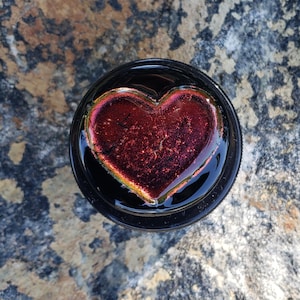 May include: A dark red glass heart with iridescent accents sits on a black background.