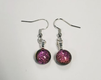 May include: A pair of dangle earrings with silver-colored hooks. Each earring features a round, dichroic glass bead with a shimmering, multi-colored effect, primarily in shades of pink and purple. The earrings are set against a white background.