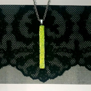 May include: A silver chain necklace with a rectangular, lime green pendant. The pendant has a textured, glittery surface. The background features a black lace pattern over a gray surface.