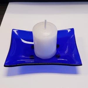 May include: A white pillar candle sits on a vibrant blue glass dish. The dish has a square shape with curved edges and a black trim. The candle is centered on the dish, creating a simple yet elegant display.