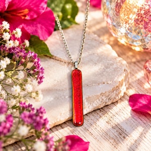 May include: A long, rectangular pendant featuring a vibrant, shimmering red glass surface with a subtle textured finish. The pendant is suspended from a simple silver-toned metal chain, creating a sleek and polished look for this piece of jewelry.