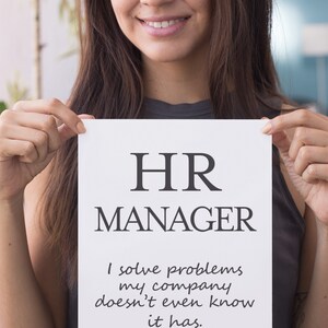 Pu&ograve; includere: Un cartello di carta bianco tenuto da una persona mostra il testo "HR MANAGER" in grassetto, nero. Sotto, il testo recita: "Risolvo problemi che la mia azienda non sa nemmeno di avere". Il cartello &egrave; un oggetto umoristico a tema ufficio.