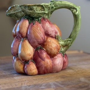 May include: A ceramic pitcher shaped like a cluster of grapes with a glossy finish. The grapes are colored in shades of purple, pink, and yellow. The handle and the top rim are sculpted to resemble green vine leaves and stems with textured details.