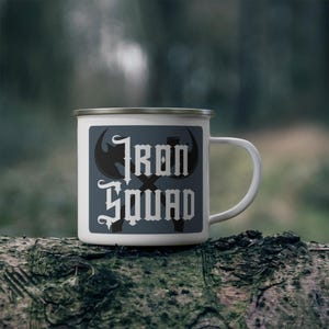 Könnte beinhalten: Weiße Emaille-Campingtasse mit einem blauen und grauen Design. Das Design zeigt eine gekreuzte Axt und den Text "Iron Squad".
