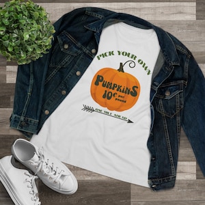 Può includere: Maglietta bianca con una freccia verde che punta a destra e il testo "Pick Your Own Pumpkins 10¢ per pound One Mile Ahead". Sopra il testo c'è una zucca arancione disegnata con uno stelo verde.
