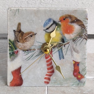 Peut inclure: Une horloge carrée en marbre blanc avec un cadran et des aiguilles dorés. Le cadran de l'horloge représente une scène d'hiver avec trois oiseaux perchés sur une branche, dont un rouge-gorge, un mésange bleue et un troglodyte brun. Les oiseaux sont perchés sur une branche avec des chaussettes rayées rouges et blanches suspendues aux branches.