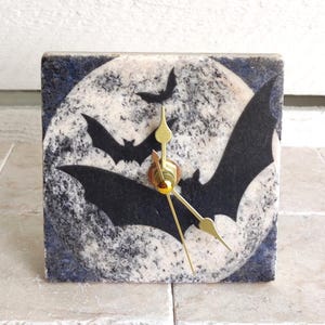 Puede incluir: Un reloj cuadrado de mármol blanco y negro con un diseño de luna y murciélagos. El reloj tiene manecillas doradas.