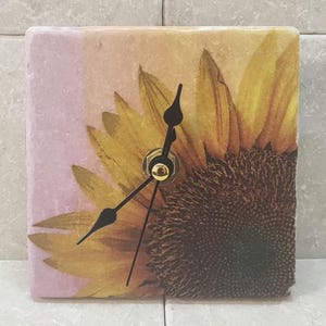 May include: A square clock with a sunflower design. The clock face is white with black hands. The sunflower is a close-up of the center of the flower with brown and yellow petals.