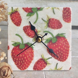 May include: A square clock with a white background and a pattern of red strawberries. The clock has black hands and a gold center.