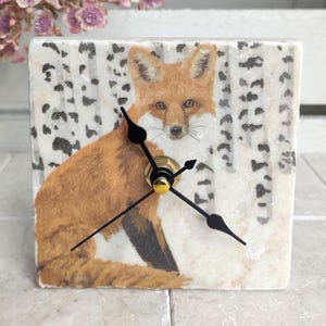 May include: A square clock with a white background and a fox illustration. The fox is brown and orange with black eyes and a black nose. The clock has black hands and a gold center. The background is a white and black birch tree pattern.