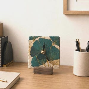 Könnte beinhalten: Eine quadratische Steinuhr mit einem Muster aus blaugrünen und goldenen Ginkgoblättern auf dem Zifferblatt. Die Uhr hat schwarze Zeiger und steht auf einem rechteckigen Sockel aus dunklem Holz auf einer ebenen, strukturierten Oberfläche.