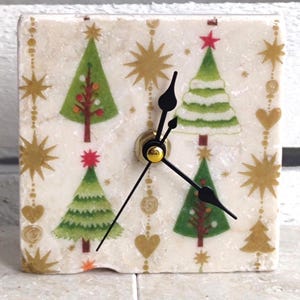 May include: White square clock with black hands and a Christmas tree design. The clock face has a white background with gold stars, hearts, and Christmas trees.