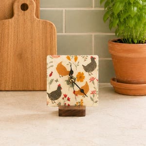 May include: A square analog clock featuring a cream-colored face decorated with illustrations of chickens, orange flowers, and red floral sprigs. The clock has black hands and is mounted on a rectangular dark brown wooden base for stability.