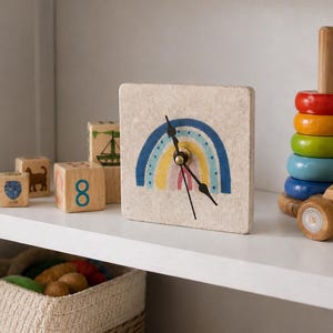 May include: A square clock with a rainbow design in blue, yellow, and pink. The clock hands are black. Wooden blocks with numbers and animal illustrations are on the shelf. A wooden stacking toy with colorful rings is also present.