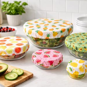 Peut inclure: Plusieurs bols en verre recouverts de couvercles alimentaires réutilisables en tissu. Les couvercles présentent des motifs de fruits colorés, notamment des ananas, des citrons, des oranges, des pastèques et des avocats. Les bols contiennent divers aliments.