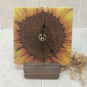 May include: A square clock with a sunflower design. The clock face is a close-up of the sunflower's center, with black hands and a gold knob. The clock is on a dark wood stand.