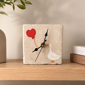 May include: A square clock with a beige stone texture features a red heart balloon and a white goose illustration. The clock hands are black, and the clock is set on a wooden shelf.