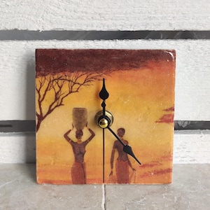 May include: A square clock with a painted image of three women walking in a desert landscape. The clock has black hands and a gold center. The background is a sunset with orange, yellow, and brown colors.