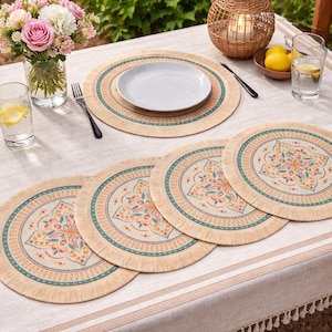 May include: A table setting with beige placemats featuring a floral design in orange, teal, and pink. The table is set with plates, glasses, cutlery, and a vase of flowers. A candle in a woven holder adds ambiance.