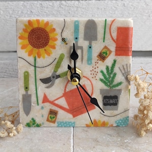 May include: A white square clock with black hands and a gold center. The clock face has a colorful pattern of gardening tools, flowers, and plants. The text "Seeds" and "Rosemary" are visible on the clock face.