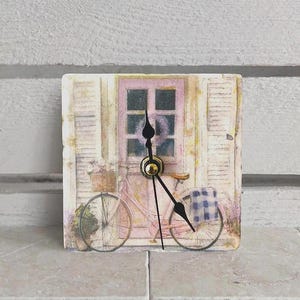 Könnte beinhalten: Quadratische Uhr mit einem Vintage-Fahrrad-Design. Das Zifferblatt zeigt ein rosa Fahrrad mit einem Blumenkorb und einem karierten Korb. Die Zeiger der Uhr sind schwarz, und die Uhr hat eine goldfarbene Mitte.