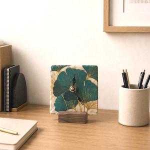 Può includere: Un orologio da scrivania quadrato con un motivo di foglie di ginkgo color verde acqua e oro sul quadrante. L'orologio presenta lancette nere e un piccolo centro dorato, ed è sostenuto da una base rettangolare in legno scuro.
