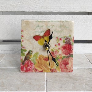 May include: A square clock with a floral design and a butterfly. The clock has black hands and a gold knob. The background is a white wall.