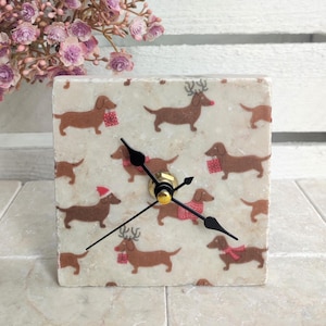 May include: Square white marble clock with black hands and a design of brown dachshunds wearing Santa hats and red scarves. Some dachshunds have reindeer antlers.