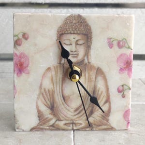 May include: A square clock with a white background and a floral design. The clock face features a painting of a meditating Buddha with closed eyes. The clock hands are black and the clock face is surrounded by pink flowers.
