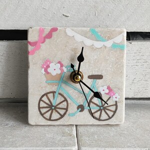 May include: Square decorative clock with a hand-painted bicycle design. The bicycle is light blue with brown wheels and a basket of pink and white flowers. The clock has black hands and a gold-coloured centre.
