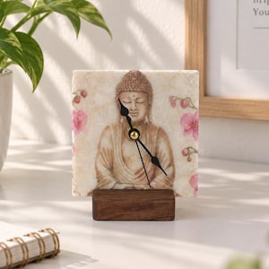 Könnte beinhalten: Eine quadratische Steinuhr mit einem aufgedruckten Bild einer sitzenden Buddha-Figur in neutralen Farbtönen, akzentuiert durch rosa Orchideenblüten an den Seiten. Die Uhr hat schwarze Zeiger und steht auf einem rechteckigen Sockel aus dunklem Holz.