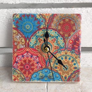 May include: A square wall clock featuring a vibrant, repeating mandala pattern in shades of red, blue, yellow, and orange. The clock face has three thin black hands centered on a gold-toned pin, displaying a colorful, intricate geometric design.