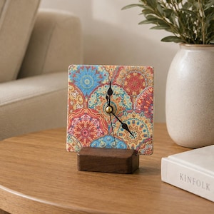 Op de afbeelding: Een vierkante analoge klok met een levendig, veelkleurig mandalapatroon in tinten rood, blauw, geel en oranje. De wijzerplaat is gemonteerd op een rechthoekige donkerbruine houten voet en heeft zwarte metalen wijzers die de tijd aangeven.