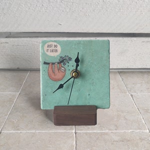 May include: A teal green clock with a sloth hanging from a branch. The clock face has black hands and a gold center. The clock is on a wooden stand. The clock face says "Just do it later".