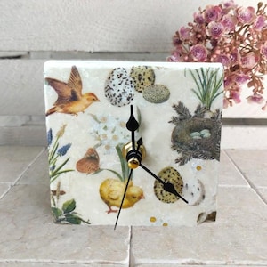 Può includere: Orologio decorativo quadrato con un design a tema uccello, farfalla, pulcino e uova. Il quadrante dell'orologio ha uno sfondo color crema con un collage a tema natura. Le lancette dell'orologio sono nere.