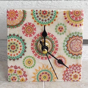 Puede incluir: Un reloj cuadrado de piedra con una base de color crema y un patrón repetitivo de coloridos diseños circulares estilo mandala en tonos rojo, verde, amarillo y naranja. El reloj tiene tres manecillas negras y un mecanismo central dorado.
