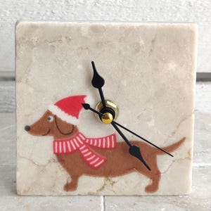 May include: A white marble clock with a dachshund wearing a red Santa hat and a red and white striped scarf. The clock hands are black.