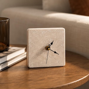 Puede incluir: Un reloj analógico cuadrado con una esfera de piedra beige texturizada. El reloj cuenta con finas manecillas negras para las horas, los minutos y los segundos, centradas en un pequeño eje circular de tono dorado. Los bordes son lisos y redondeados.