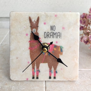 May include: A white marble clock with a llama illustration and the text "NO DRAMA!" in black lettering. The llama is wearing a pink and blue patterned saddle and has pink and blue leg warmers.