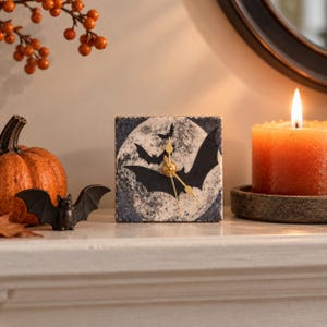 May include: A square analog clock featuring a graphic of black bats flying in front of a full moon. The clock face has a textured, mottled gray and white background with gold-colored hour, minute, and second hands centered on the design.
