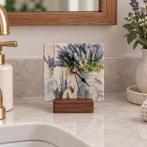 Puede incluir: Un reloj de mesa cuadrado con una ilustración impresa de ramas de lavanda en una botella de vidrio y un frasco con un motivo de abeja. La esfera está montada sobre una base rectangular de madera oscura y tiene manecillas negras.