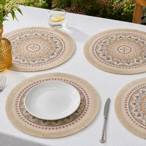 Op de afbeelding: Vier beige ronde placemats met een gedetailleerd bloemenpatroon in oranje, rood en blauw. Elke mat heeft een gefranjerde rand. Een wit bord, een vork en een mes liggen op tafel, samen met een gouden vaas met bloemen en een glas water.