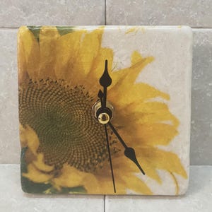 May include: Square clock with a sunflower design. The clock face is white with black hands and a gold center. The sunflower is yellow with a brown center.