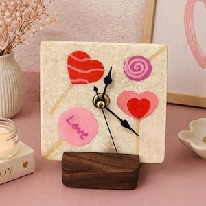 May include: A square clock with a cream-colored face featuring red and pink heart and swirl lollipop designs. The clock has black hands, a gold-colored center, and a dark wood base. The word "Love" is written on a pink circle.