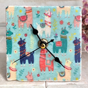 Könnte beinhalten: Eine quadratische Uhr mit blauem Hintergrund und einem Muster aus bunten Lamas. Die Uhr hat schwarze Zeiger und eine goldene Mitte.