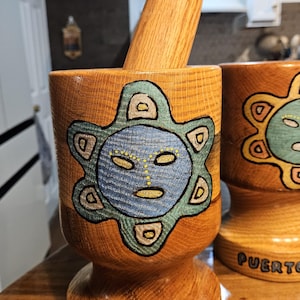 May include: A wooden mortar and pestle set featuring a carved and painted sun-like face design in blue, green, and yellow. The base of the mortar is inscribed with the text PUERTO RICO in black lettering, showcasing the natural wood grain texture.