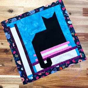 May include: A handmade quilt featuring a black cat silhouette against a blue background. The quilt has a floral border with pink, white, and purple accents. The cat is sitting on a striped platform.