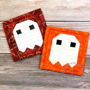 May include: Two square fabric coasters with ghost designs. One coaster has a white ghost on a red background with gold accents. The other has a white ghost on an orange background. The ghosts have black square eyes and jagged mouths.