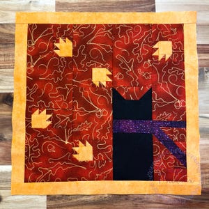 May include: A square fabric quilt block featuring a dark red background with a white outline of oak leaves and acorns. A solid black cat silhouette sits on the right side wearing a purple polka dot scarf, accented by four yellow paw prints.